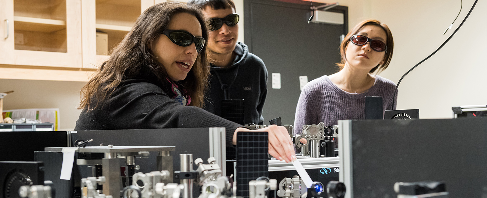 Two students look on as a professor indicates a laser beam with a pointer.