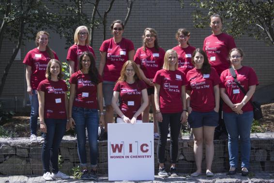 Members of the Women in Chemistry group smile for a photo with a sign bearing their logo.