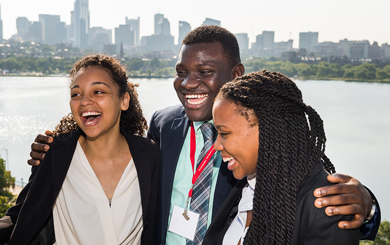 A trio of students of color smile in front of the Boston skyline.