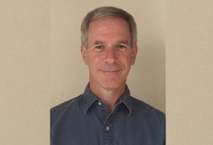 A man smiles in front of a tan wall