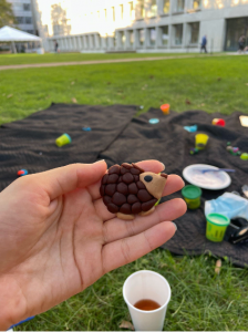 A hand holds a small hedgehog made of clay. In the background is an outdoor craft setup of others fashioning animals out of clay.