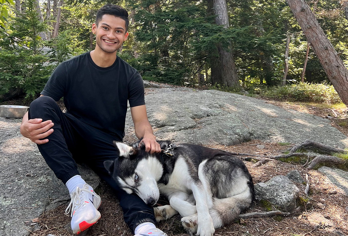 David Sarabia smiles on a mountain trail with a husky laying beside him.