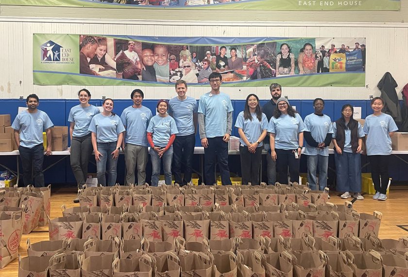 A group of volunteers wearing light blue t-shirts smiles in a row in front of dozens of empty paper bags that they are going to fill with food donations.