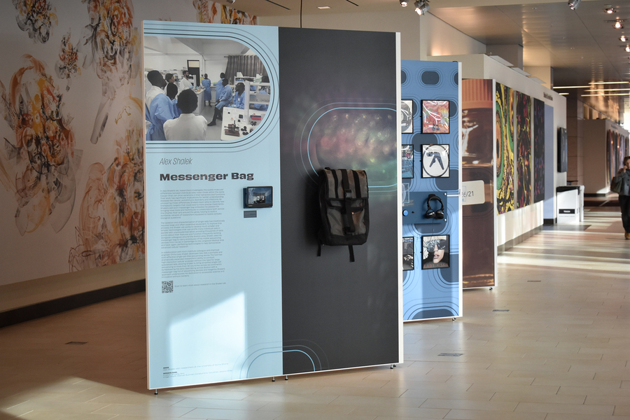 A exhibit panel in a gallery displaying a photo of numerous scientists in a lab as well as a small device and a large grey backpack.